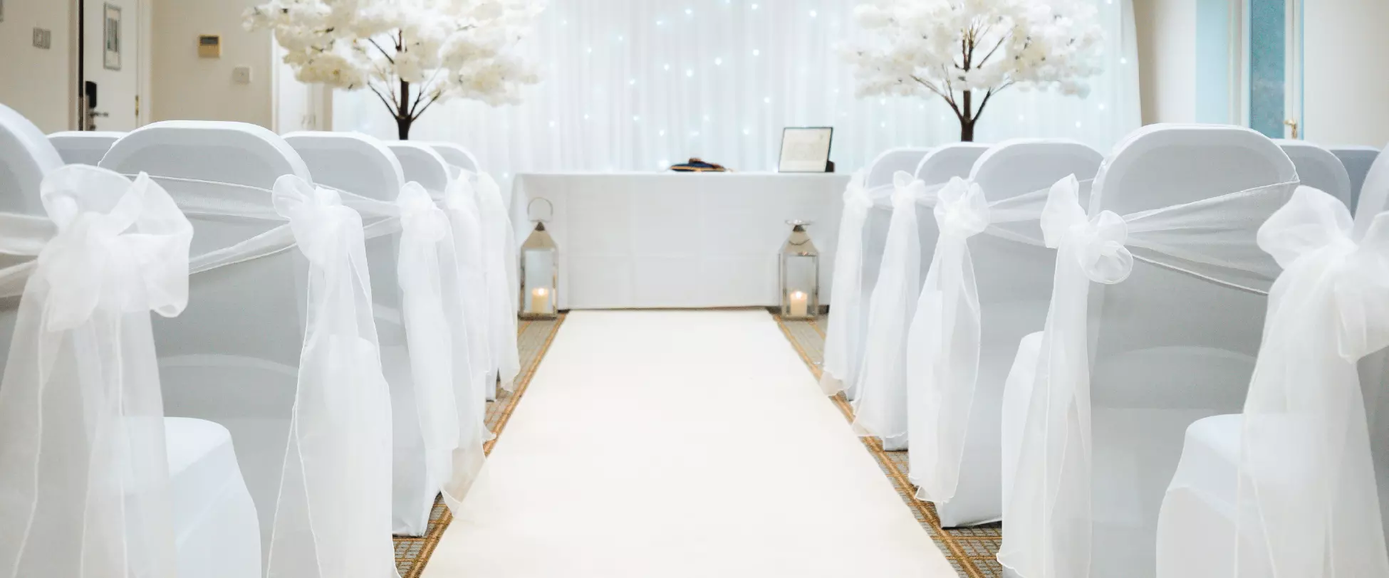 A wedding isle lined with chairs in white ribbons. there's a white carpet running up to a white table in front of a backdrop of white flowing cloth. There are two trees with white flowers, one on either side of the table..